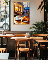 A framed art print depicting a cozy cafe scene with a croissant and coffee cup, displayed on a wall in a modern, sunlit cafe interior with wooden tables and chairs.