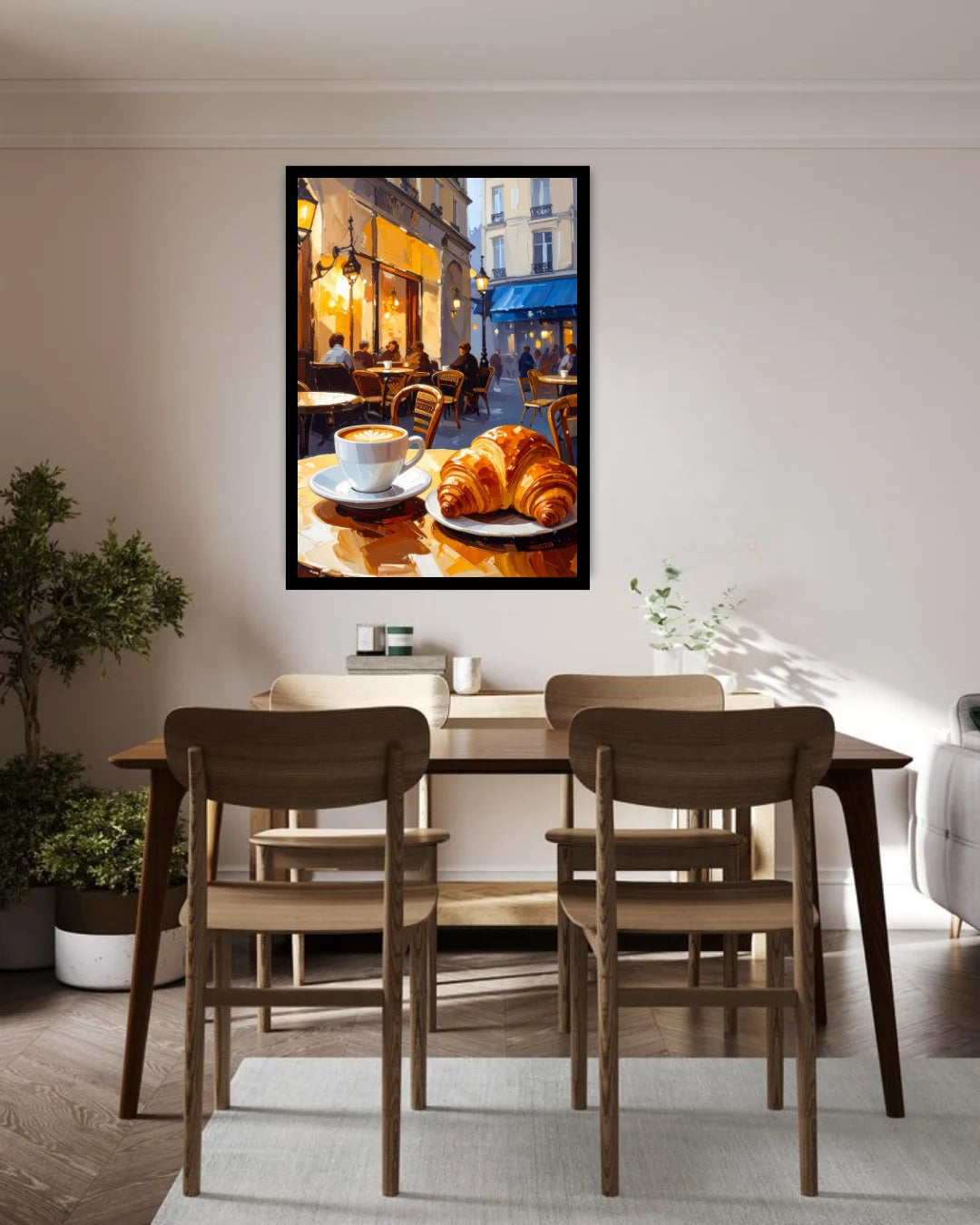 A framed wall art print depicting a cozy cafe street scene with croissants and a coffee cup, displayed above a wooden dining table and chairs in a modern, sunlit room.