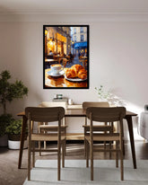A framed wall art print depicting a cozy cafe street scene with croissants and a coffee cup, displayed above a wooden dining table and chairs in a modern, sunlit room.