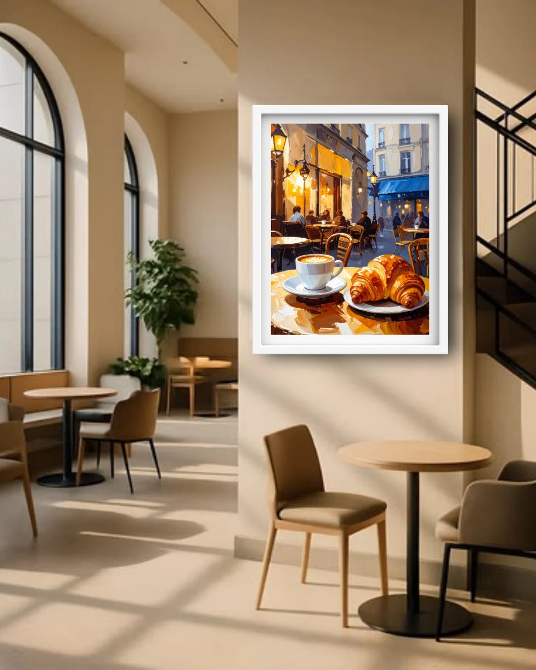 A framed wall art print depicting a cozy cafe scene with a cup of coffee and croissants, displayed on a beige wall in a modern, sunlit cafe interior.