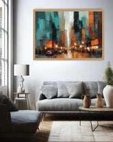 A framed abstract cityscape painting hangs on a white wall above a gray sofa in a modern living room. The artwork features teal, orange, black, and cream tones with glowing streetlights and tall buildings.