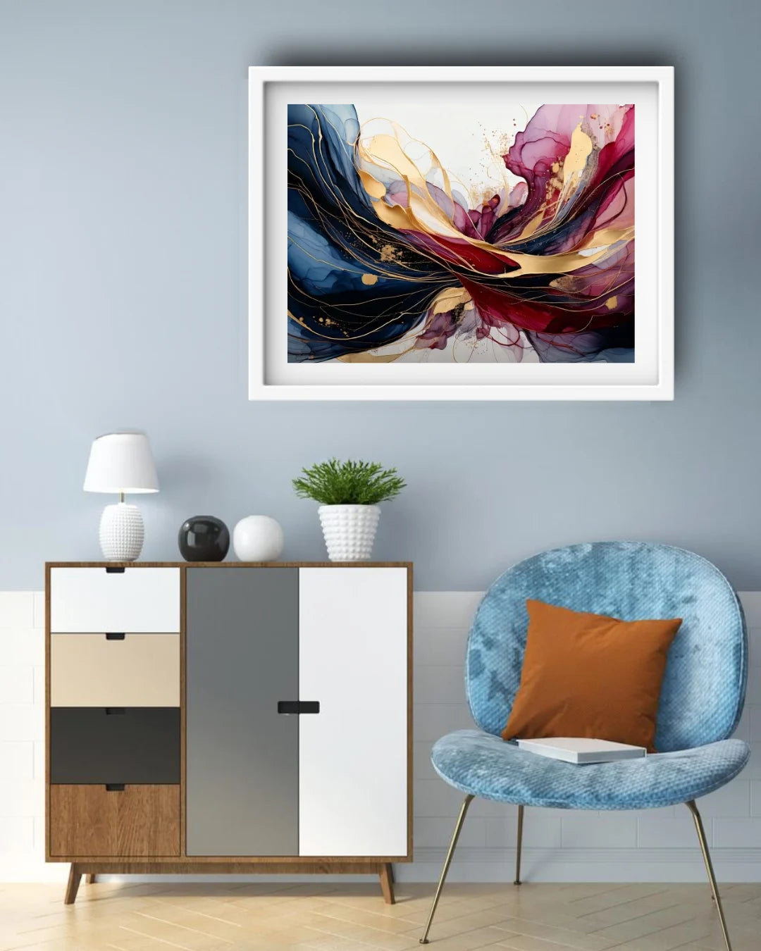 A framed abstract wall art print hangs on a light blue-gray wall above a modern cabinet, with a blue chair and orange pillow in the room. The artwork features flowing brushstrokes in navy, burgundy, gold, cream, and white.
