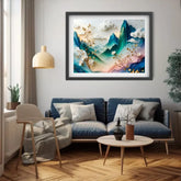 A framed abstract landscape art print hangs on a light gray wall above a blue sofa in a modern living room. The artwork features layered mountains in teal, blue, and pink tones with white clouds and golden branches.