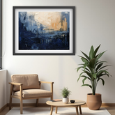 A framed abstract painting with deep blue, navy, and beige brushstrokes, displayed on a light gray wall in a modern living room setting with a beige armchair, a small wooden coffee table, and a potted green plant.