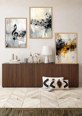 Three framed abstract wall art prints with musical notes hang above a brown ribbed console table in a styled room. The prints feature black, white, gray, and yellow-orange brushstrokes, with a white table lamp and decorative pillows on the table.