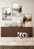 Three framed abstract wall art prints with musical notes hang above a brown ribbed console table in a styled room. The prints feature black, white, gray, and yellow-orange brushstrokes, with a white table lamp and decorative pillows on the table.