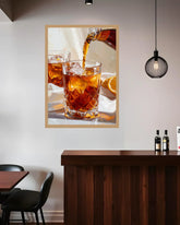 A framed wall art print depicting an amber drink being poured into a faceted glass, displayed above a dark wood counter in a modern interior setting.