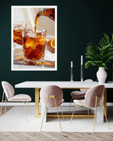 A framed wall art print showing amber liquor being poured into a faceted glass, displayed above a modern dining table with blush pink velvet chairs and gold legs against a dark green wall.
