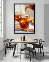 A framed wall art print depicting an amber drink being poured into a faceted glass, displayed above a dining table with chairs in a modern interior.