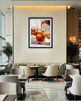 A framed wall art print depicting an iced drink with amber liquid and ice, displayed in a modern restaurant or lounge setting with tables, chairs, and warm ambient lighting.