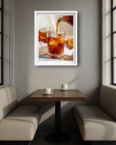 A framed wall art print depicting amber liquid being poured into an iced glass, displayed above a small booth table in a modern cafe interior.