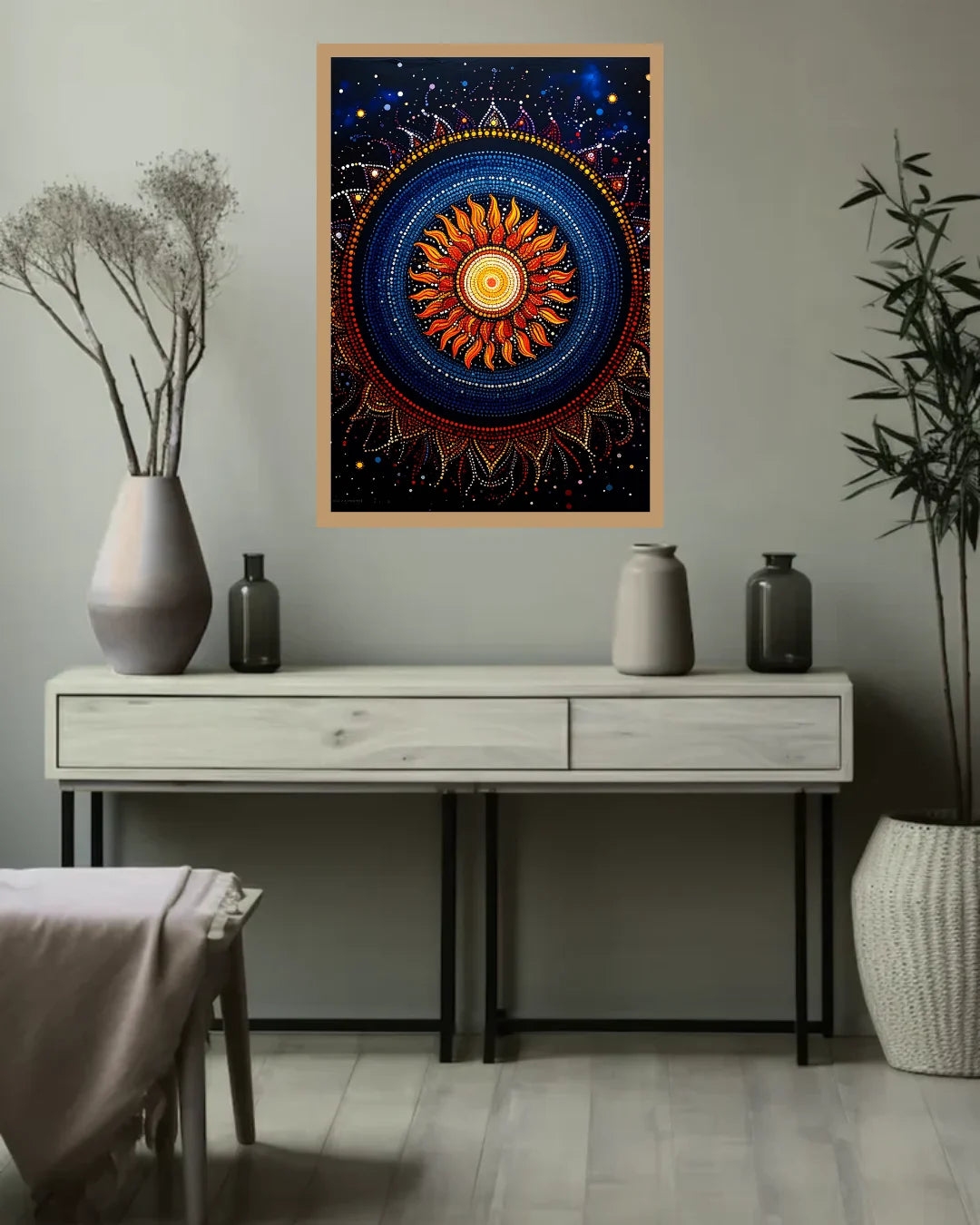 A framed square wall art print featuring a colorful mandala-style sun design hangs above a console table in a styled room with vases, bottles, and a plant.