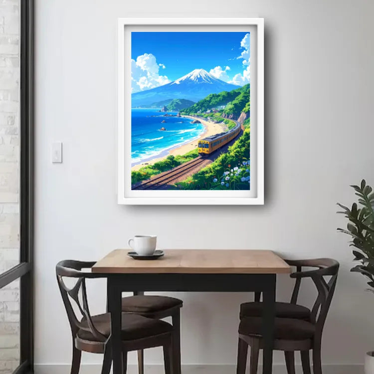 A framed art print depicting a scenic Japanese coastal landscape with a train, beach, ocean, and a snow-capped mountain, displayed on a light gray wall above a wooden dining table and chairs.