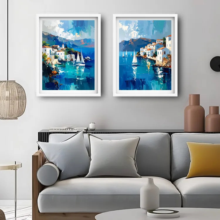 Two framed coastal paintings with blue water, white buildings, and sailboats displayed side by side above a light gray sofa in a modern living room.