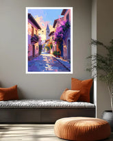 A vertical framed art print depicting a colorful, impressionistic European street scene with purple flowers and a church tower, displayed on a gray wall above a modern daybed in a styled living room.