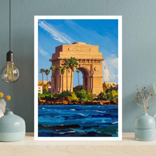 A vertical framed art print depicting a colorful painting of the India Gate in Kolkata, featuring a golden arch, palm trees, and a blue sea in the foreground, styled on a light wood table against a blue-gray wall with decorative objects.