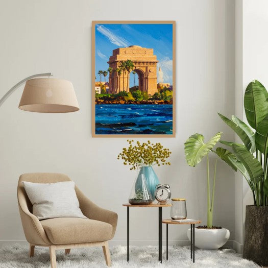 A framed painting of the India Gate monument in Kolkata, displayed on a light gray wall in a modern living room setting with a beige armchair, side tables, a lamp, and potted plants.