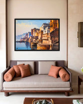 A framed painting of a colorful Indian ghats and river scene hangs on a beige wall above a modern sofa with brown and terracotta cushions in a styled living room.