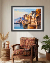A framed painting of a Ganga riverfront city scene with orange and yellow buildings, blue water, and a pale sky, displayed on a beige wall above a patterned wooden armchair in a styled room.