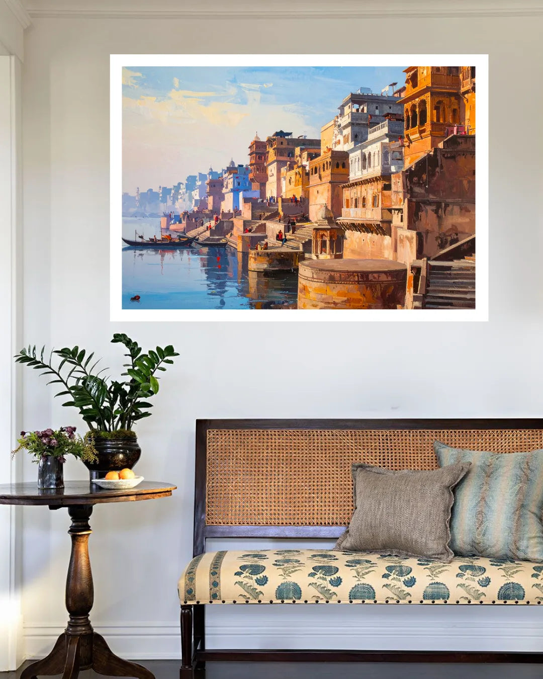 A framed art print depicting a scenic Ganga riverfront with golden buildings and boats, displayed on a white wall above a woven-back bench in a styled room.