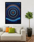 A framed abstract wall art print featuring concentric circular and wavy dot patterns in deep blue, navy, black, white, and small gold accents, displayed above a light beige sofa in a modern living room setting with decorative pillows and a potted tree.
