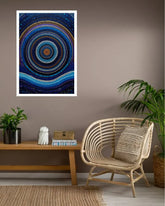 A framed abstract wall art print featuring concentric circles and flowing wave-like bands in deep blue, teal, and white, displayed on a taupe wall above a wooden bench with plants and a rattan chair in a styled room.