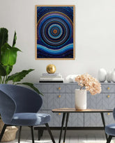 A framed abstract art print featuring concentric circles and dots in shades of blue, navy, and gold, displayed on a light gray wall in a modern living room setting.