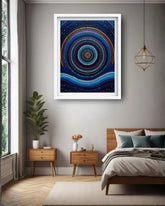 A framed abstract art print featuring concentric circles and dots in shades of blue, black, and gold, displayed on a gray wall in a modern bedroom setting.