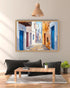 A framed painting of a narrow street with blue doors and white and ochre walls, displayed on a beige wall above a wooden coffee table and black cushions in a modern living room setting.