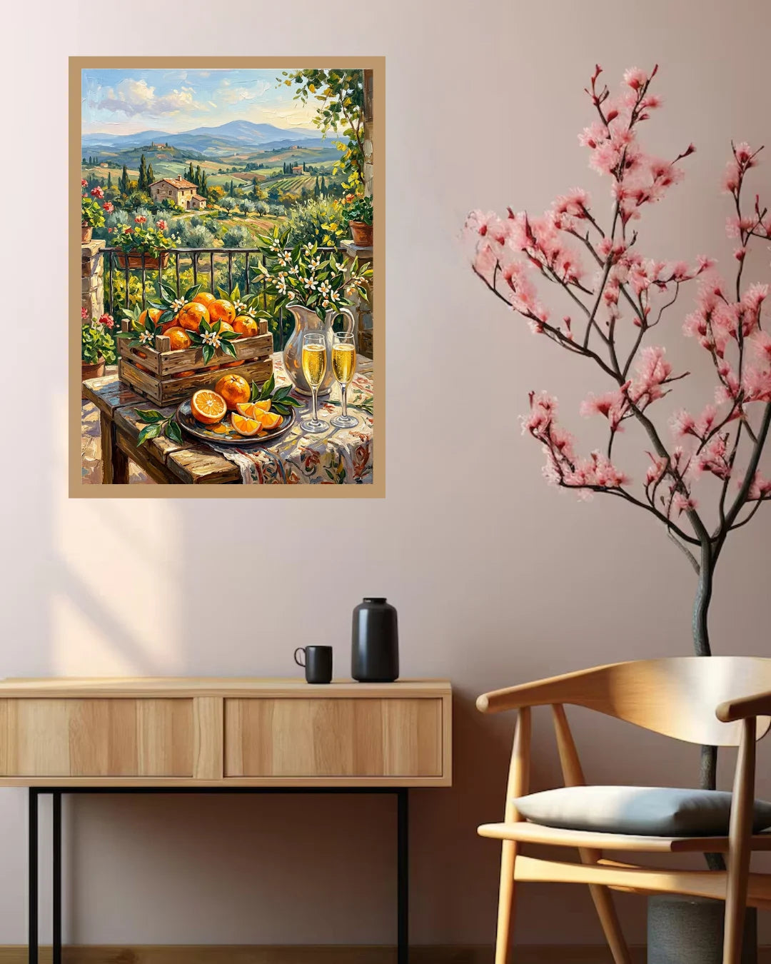 A framed art print depicting a scenic Italian landscape with a still life of oranges and glasses of white wine, displayed on a beige wall above a light wood console table and next to a wooden chair with a pink flowering branch decoration.