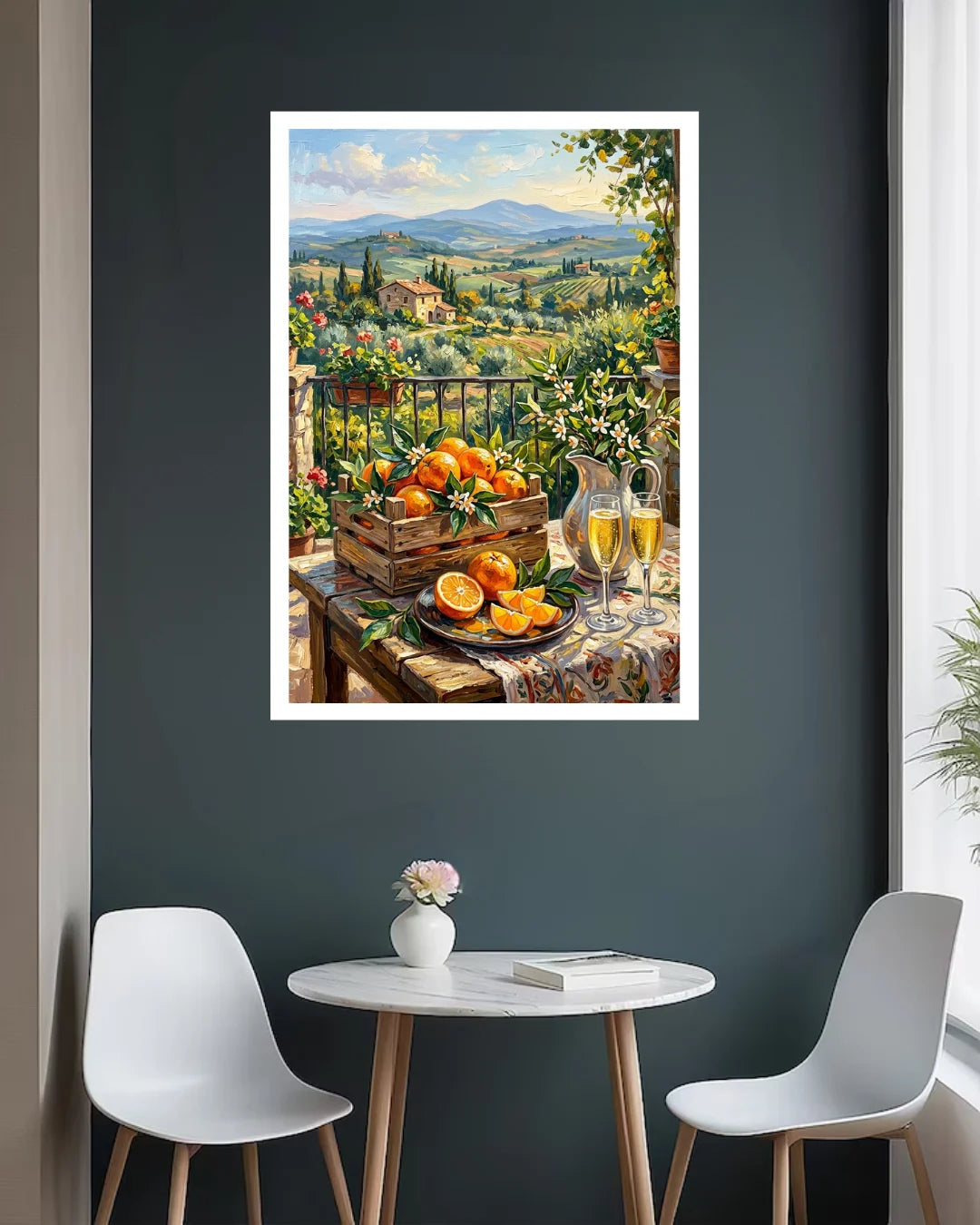 A framed art print depicting a vibrant Italian landscape with oranges on a table, hung on a dark gray wall above a small white table and two modern chairs.