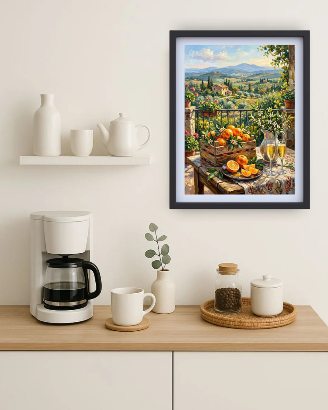 A framed art print depicting a scenic Italian landscape with a fruit table and wine glasses, styled on a light wood kitchen counter next to a coffee maker and white ceramic mugs.