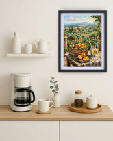 A framed art print depicting a scenic Italian landscape with a fruit table and wine glasses, styled on a light wood kitchen counter next to a coffee maker and white ceramic mugs.