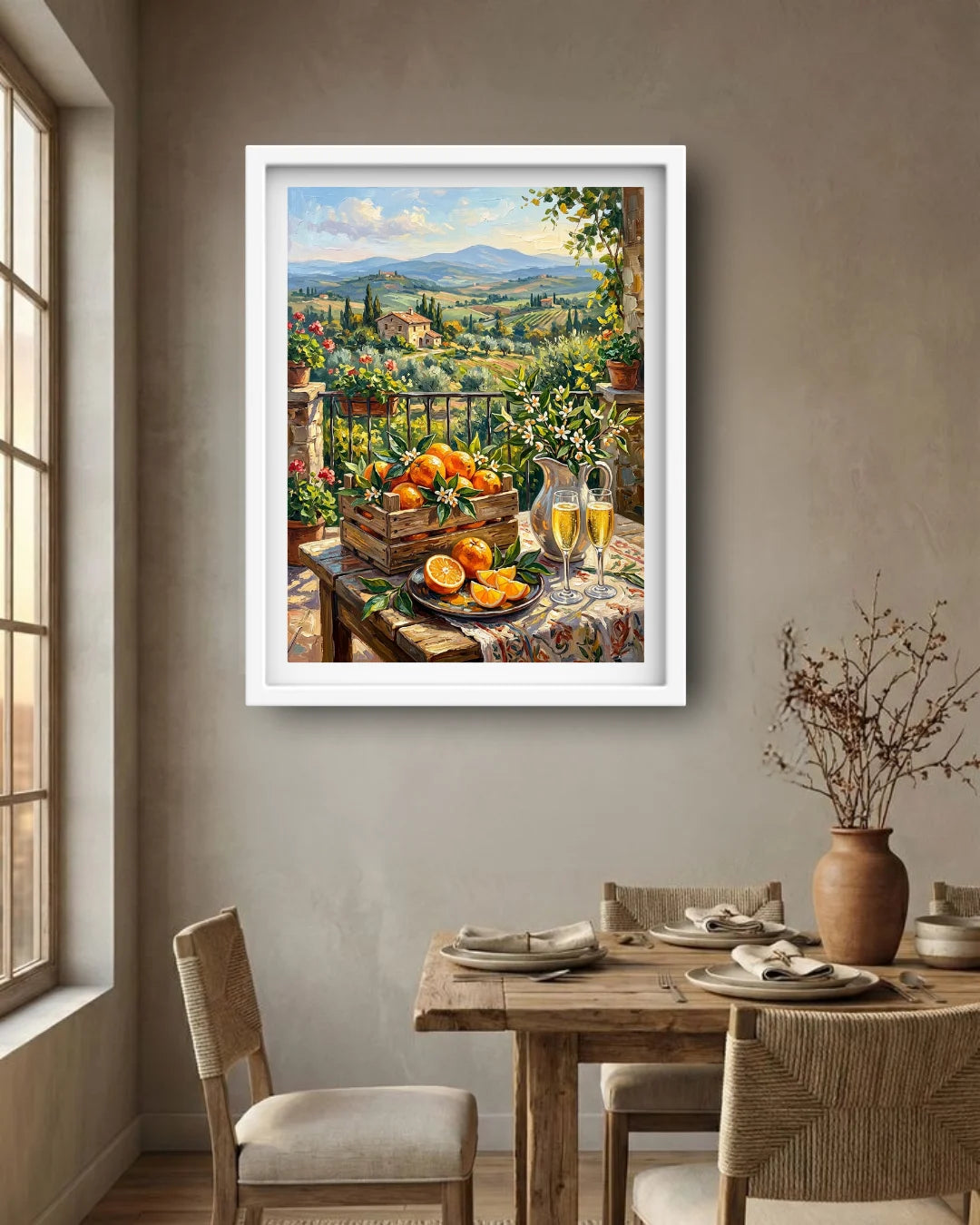 A framed landscape painting of a Tuscan vineyard and fruit still life hangs on a beige wall above a wooden dining table set with chairs and a vase in a dining room.