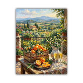 A vertical canvas art print depicting a sunny Tuscan landscape with rolling hills, cypress trees, and a stone villa, styled with a wooden crate of oranges, a pitcher, and two glasses of white wine on a table in the foreground.