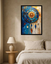 A framed abstract dreamcatcher-style wall art print hangs above a bed in a softly lit bedroom. The artwork features a circular web motif with blue, gold, orange, and white tones, and dangling jewel-like shapes.