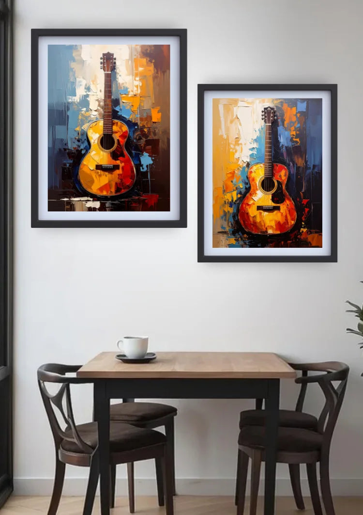 Two framed abstract paintings of acoustic guitars in warm orange, yellow, and blue tones, displayed on a white wall above a wooden dining table with two chairs.