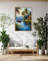 A framed tropical landscape painting hangs on a white wall above a light wood sofa with white cushions in a bright, modern living room. The painting depicts palm trees, a small boat on blue water, and a house, surrounded by potted green plants.