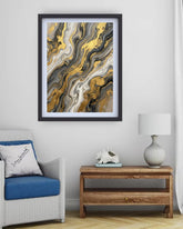 A framed abstract art print with swirling gold, black, gray, and white marble-like patterns, displayed on a light gray wall above a wooden side table and next to a white armchair with a blue cushion.