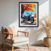 A framed vertical art print depicting a blue motorcycle under a vibrant orange autumn tree, displayed on a white wall in a styled living room setting with a beige armchair and a potted pampas grass plant.