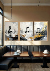 Three framed abstract wall art prints with musical notes and staff lines hang above a black leather booth in a cafe with wooden tables and coffee cups.