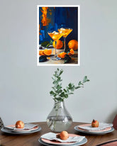A framed art print depicting two orange Martini glasses with oranges on a table, displayed on a light gray wall above a dining table set with plates, napkins, and a glass vase.