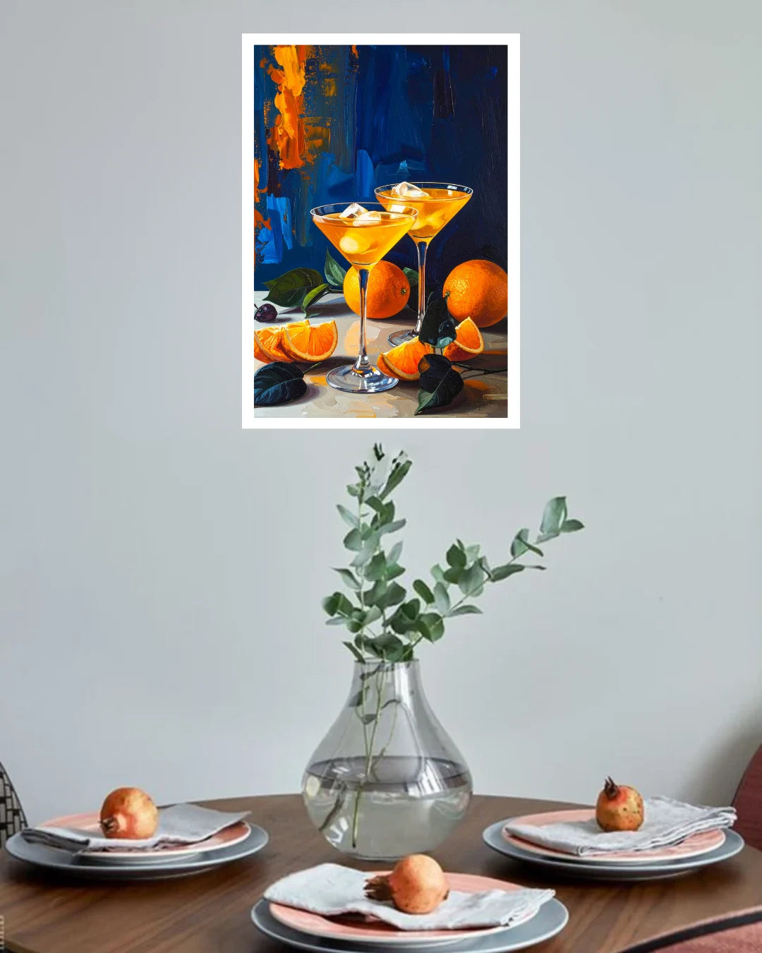 A framed art print depicting two orange Martini glasses with oranges on a table, displayed on a light gray wall above a dining table set with plates, napkins, and a glass vase.