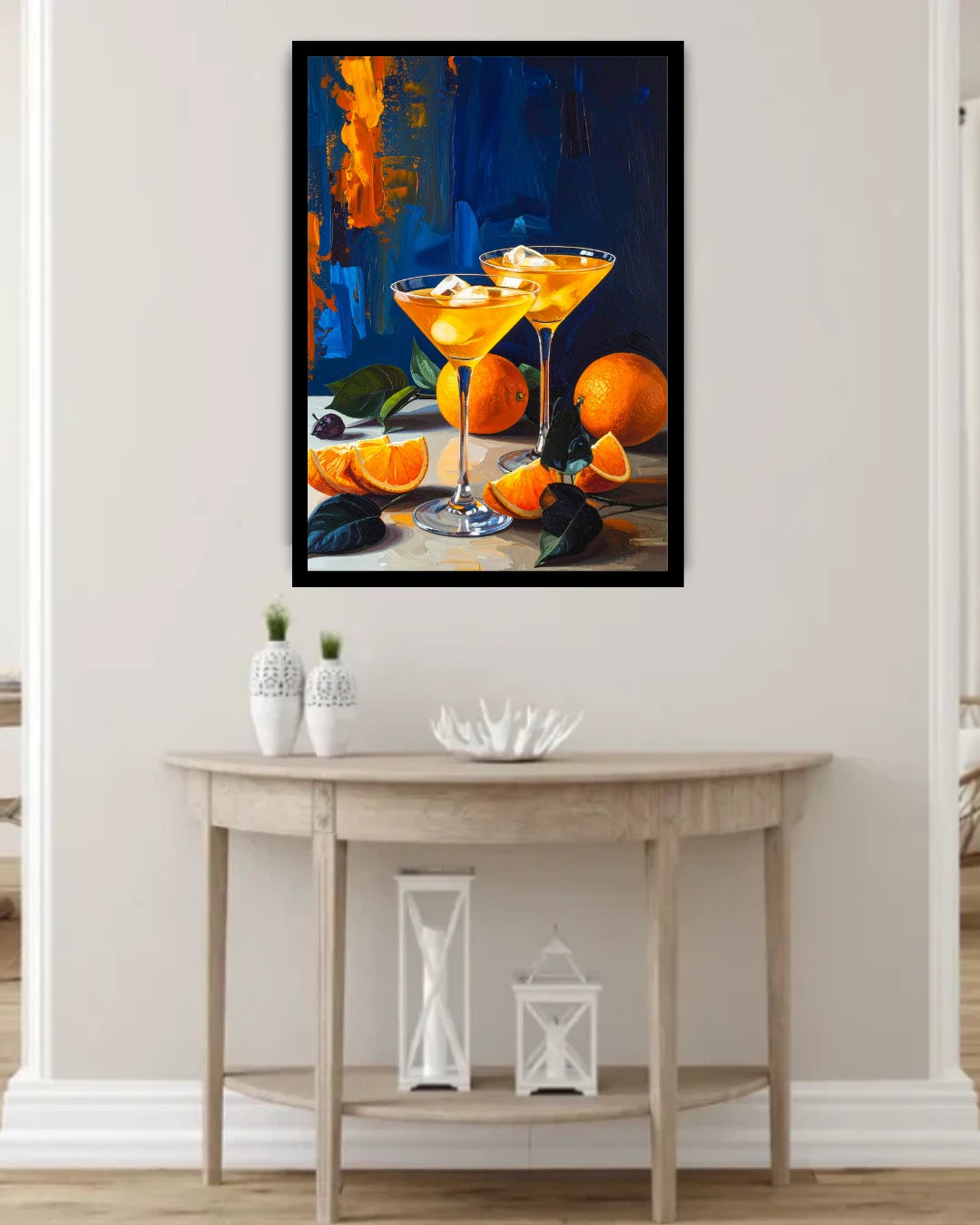 A framed vertical canvas print of a still life painting featuring two orange cocktails and oranges, displayed on a beige wall above a light wood console table with decorative vases and lanterns.