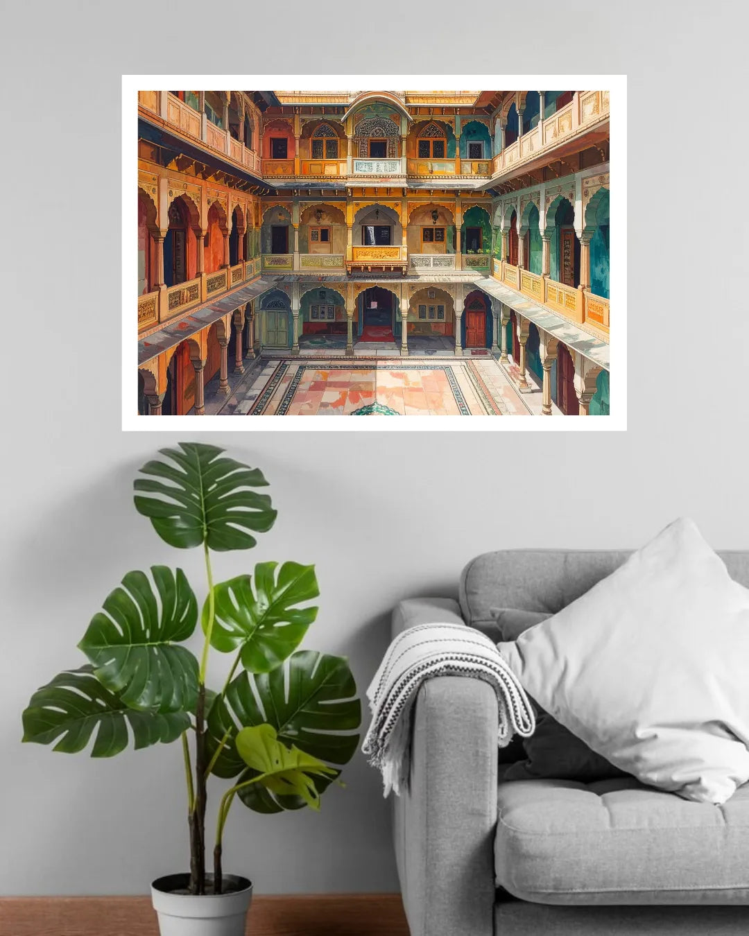 A framed art print depicting a colorful traditional courtyard with arches and balconies, displayed on a light gray wall above a gray sofa with a white pillow and a potted green plant.