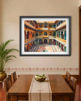 A framed art print depicting an ornate Indian courtyard with arches and balconies, displayed on a beige wall above a wooden dining table with chairs.