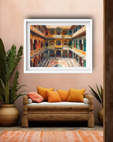 A framed art print depicting a colorful courtyard in a traditional Indian palace, displayed on a beige wall above a rattan sofa with orange cushions in a styled room.