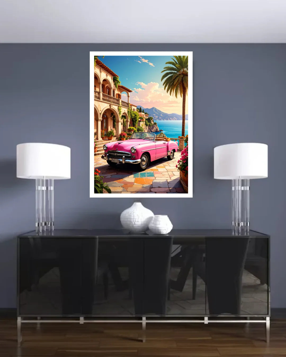 A framed art print depicting a classic pink convertible car parked on a colorful tiled street by a coastal villa with palm trees and blue water, displayed on a dark gray wall above a black console table with two white table lamps.
