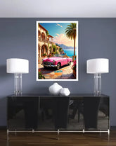 A framed art print depicting a classic pink convertible car parked on a colorful tiled street by a coastal villa with palm trees and blue water, displayed on a dark gray wall above a black console table with two white table lamps.