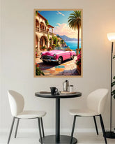 A framed art print depicting a vibrant pink convertible car parked in front of a Mediterranean-style building with a palm tree and blue sea, displayed on a wall above a small dining table with two chairs.